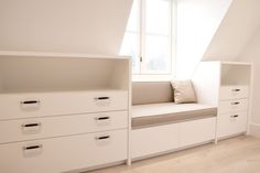 a white room with drawers and a window in the corner next to a bench that has a cushion on it