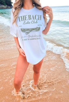 a beautiful woman standing on top of a sandy beach next to the ocean wearing a white shirt