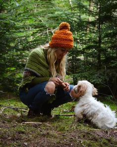 a woman kneeling down next to a dog in the woods with a knitted hat on