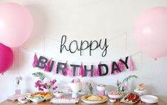 a birthday party with pink balloons and food on a wooden table in front of a sign that says happy birthday