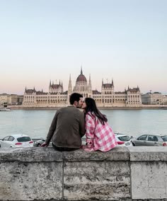 a man and woman are sitting on a stone wall looking at the water in front of a large building