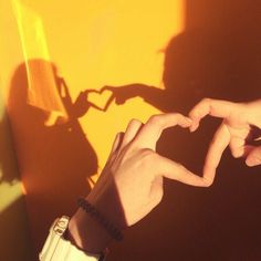 two hands making a heart shape with their fingers against a yellow wall and shadows cast on the wall