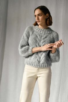 a woman standing in front of a white backdrop wearing a gray sweater and cream pants