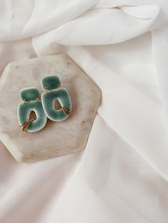 a pair of green earrings sitting on top of a white cloth covered tablecloth with an object in the background