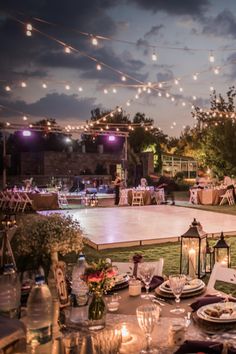 an outdoor dinner setup with lights strung over the tables and in the distance, there is a lit dance floor