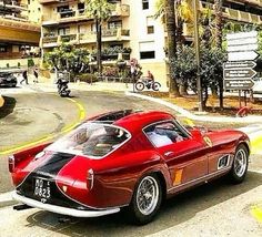 an old red sports car driving down the street in front of tall buildings and palm trees