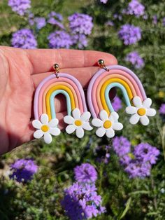 a pair of rainbow earrings with daisies in front of purple flowers