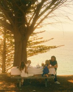 a group of people sitting at a picnic table under a tree near the water's edge