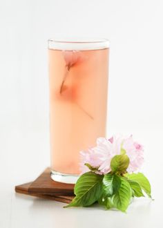 a pink drink in a tall glass next to some flowers on a wooden board with green leaves