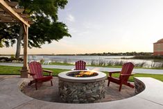 two red chairs sitting next to a fire pit on top of a stone patio near a lake