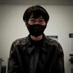 a man wearing a black mask and leather jacket in an office setting with papers on the wall behind him