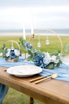 a wooden table topped with blue flowers and white plates covered in greenery next to candles