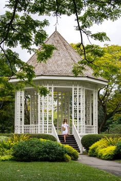 a woman standing in front of a white gazebo surrounded by trees and bushes on a sunny day