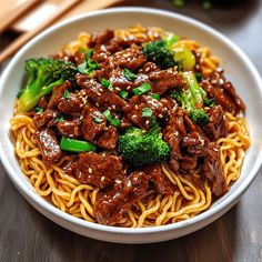 a white bowl filled with beef and broccoli on top of noodles next to chopsticks