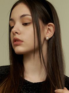 a woman with long brown hair wearing a black shirt and pearled earrings, looking down at her left ear