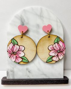 two wooden earrings with pink flowers and hearts on them sitting on a white marble surface