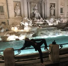 a woman leaning on a railing in front of a fountain with statues around it at night