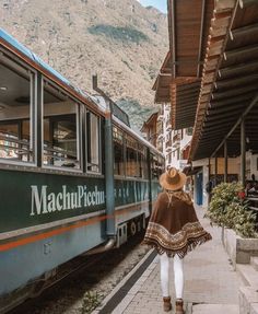 a woman walking down the sidewalk next to a train at a train station with mountains in the background