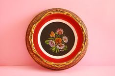 a red and gold plate with flowers painted on the side is against a pink background