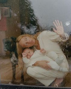 a woman holding a baby in front of a window with a dog looking on from behind it