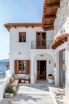 a white house with brown shutters and wooden doors on the front porch overlooking the water