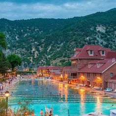 people are swimming in a large pool at the foot of a mountain side town with mountains in the background