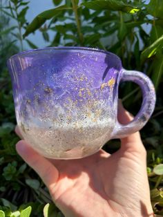 a hand holding a purple and white coffee cup in front of some green plants with yellow speckles on it