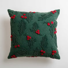 a green pillow with red berries and leaves embroidered on the front, sitting against a white wall