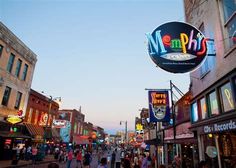 people are walking down the street in front of shops and businesses at dusk, with neon signs above them