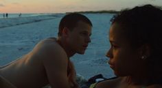 a man and woman laying on the beach at sunset looking into each other's eyes