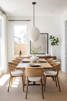 a dining room table with white chairs and a vase on top of it, in front of large windows