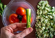 a person holding up a piece of cucumber next to a bowl with tomatoes, celery and other vegetables