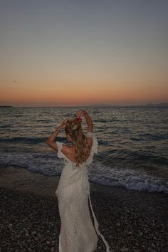 a woman standing on top of a beach next to the ocean at sunset with her hair blowing in the wind