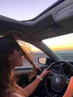 a woman driving a car with the sun setting in the background and water behind her