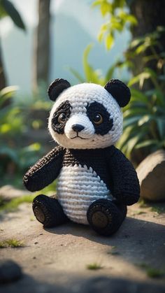 a crocheted panda bear sitting on the ground in front of some trees and bushes