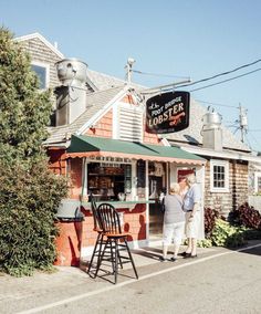 an older couple standing outside of a small store on the side of the road in front of a house