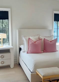 a white bed sitting next to two windows with pink pillows on top of each pillow