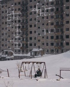 a man sitting on top of a swing set in the snow next to a tall building