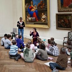 a group of children sitting on the floor in front of a painting with a woman standing next to them