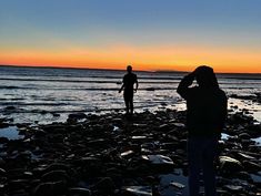 two people are standing on the beach watching the sun go down over the water and rocks