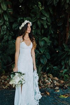 a woman in a white dress holding a bouquet and wearing a flower headpiece standing next to trees