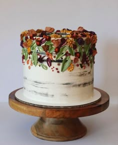 a white cake decorated with flowers and leaves on top of a wooden stand in front of a wall