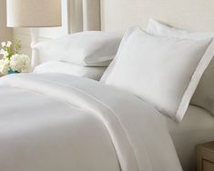 a bed with white sheets and pillows on top of it next to a nightstand with flowers