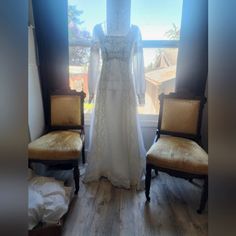 a wedding dress is displayed in front of a window with an old chair next to it