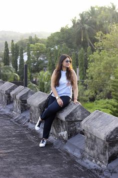 a woman sitting on the edge of a stone wall with trees and bushes in the background