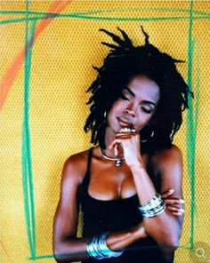 a woman with dreadlocks standing in front of a yellow and green background holding her hand to her chin