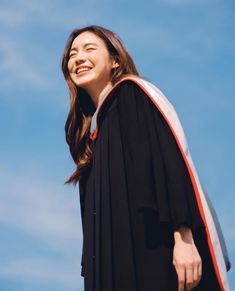 a woman wearing a black graduation gown and smiling at the camera while standing in front of a blue sky