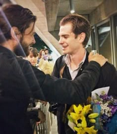 two men shaking hands in front of a group of people with flowers on the street