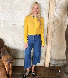 a woman standing in front of a wall wearing jeans and a yellow shirt with buttons on it