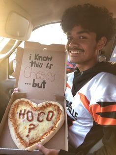 a man holding up a heart shaped pizza in the back seat of a car with an i know this is cheesy but hoco written on it